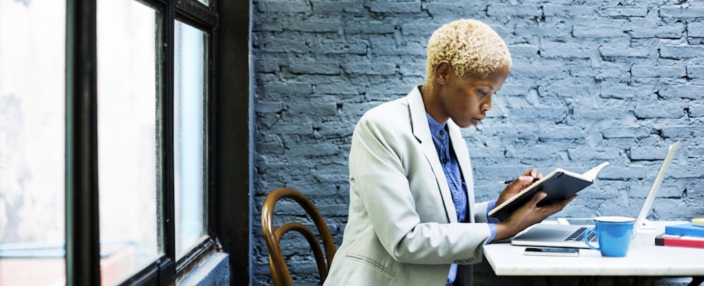 Woman sitting in a gray room with a book and laptop to learn about the ideal debt to income ration.