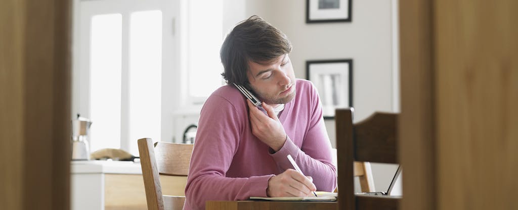 Man sitting in kitchen, speaking on phone with his credit card company and negotiating his debt.