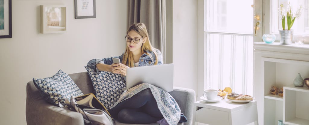 Young women relaxing on a couch in a sunny room, looking at her phone with her laptop on her lap