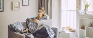 Young women relaxing on a couch in a sunny room, looking at her phone with her laptop on her lap