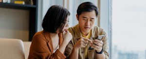 A couple seated on a sofa use a mobile phone to close a bank account online.