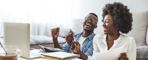 A seated couple laugh as they use a laptop and calculator to help them determine what their home is worth.