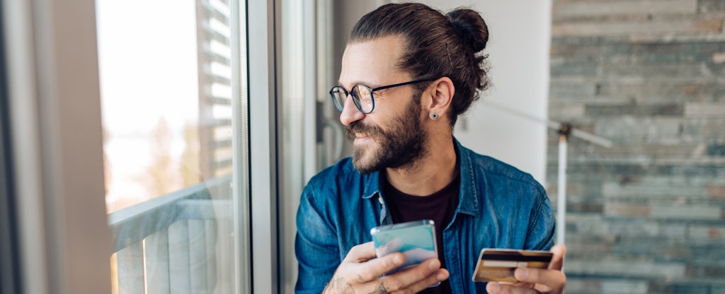 A smiling man looks out the window while holding his smartphone and credit card.