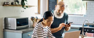 A couple in their kitchen use a smartphone and a laptop to learn how to deposit a check online with their bank.