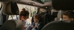 A mother buckles a small child into a carseat in the backseat of a car while another child looks on.