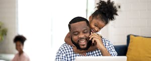 Man working on his laptop while his child throws her arms around him, asking him to play