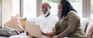 A smiling couple seated on a sofa hold a laptop while looking into rates for a Navy Federal HELOC.