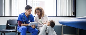 A nurse in a hospital room shows a digital tablet to a parent whose small toddler leans against them.