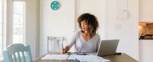A person seated at their dining room table uses laptop and financial documents to calculate their net income