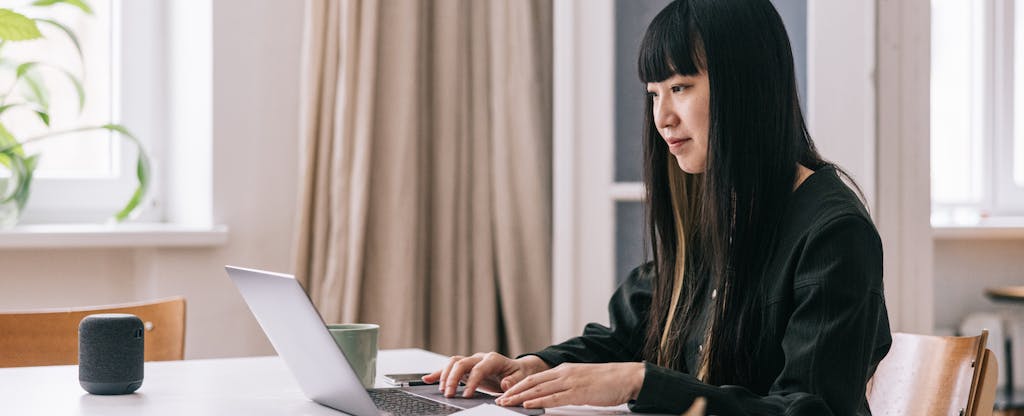 A person with long hair sits at their dining room table and uses their laptop to research how to make a financial plan