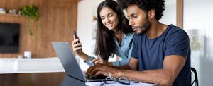 A couple seated at a table use a laptop and mobile phone while filing their taxes online.